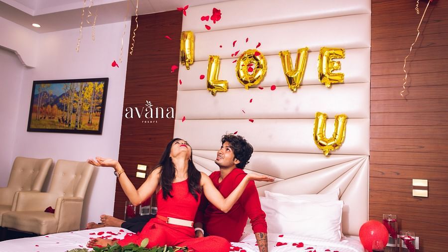 A couple sitting on a bed decorated with rose petals and  I LOVE U balloons.