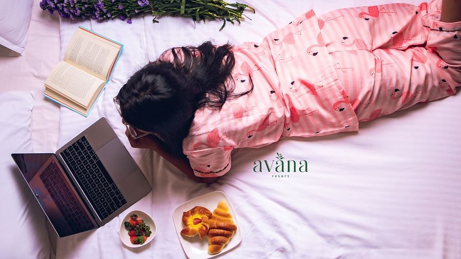 Woman in night dress relaxing on bed with breakfast and laptop, surrounded by books and flowers.
