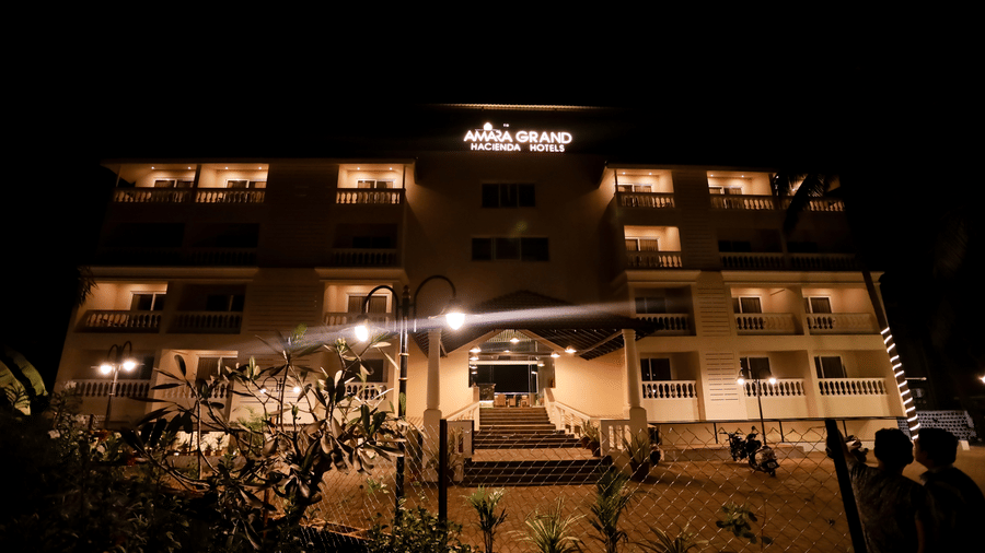 Night view of Amara Grand Baga, Goa, with a row of plants in front of it.