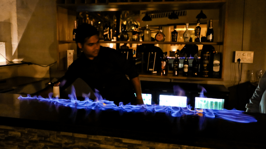 A bar with a man standing behind the counter with drinks behind him and a flame across the table at Amara Grand Baga, Goa.