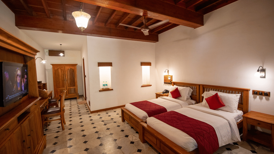 A spacious bedroom at Chidambara Vilas - A Luxury Heritage Resort, with a twin beds, tiled flooring, wooden beams on the ceiling, and natural light from a window.