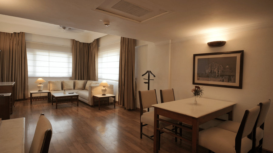 A large living and dining area with wooden flooring, a dining table, upholstered chairs, and wide windows allowing ample natural light in the Executive Suite at Clarks Amer, Jaipur.