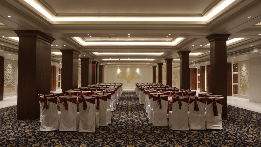 An elegant hotel function room features rows of chairs with white covers and dark red sashes set on patterned carpet - Clarks Amer, Jaipur