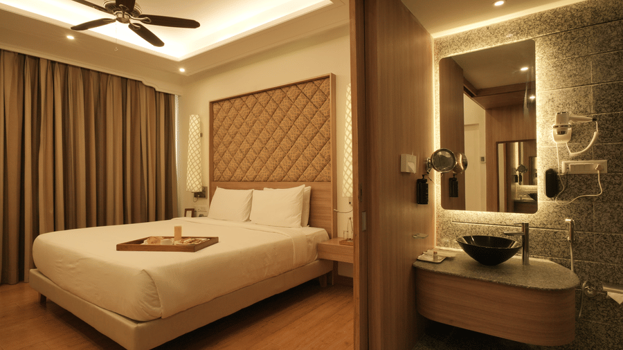 A spacious bedroom with a king size bed, upholstered headboard, wooden flooring, and an attached open bathroom area with a glass partition in the Presidential Suite at Clarks Amer, Jaipur.
