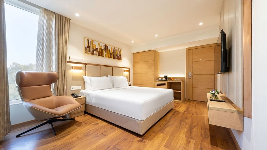 A spacious hotel room with a large bed, a comfortable armchair, a flat-screen TV, and a large window with curtains - Clarks Inn, Indore