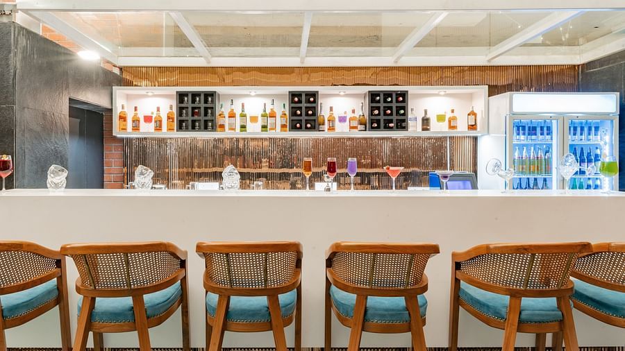 A bar with a white counter, a row of wooden stools with blue cushions, and a shelf displaying bottles of liquor - Clarks Inn, Indore