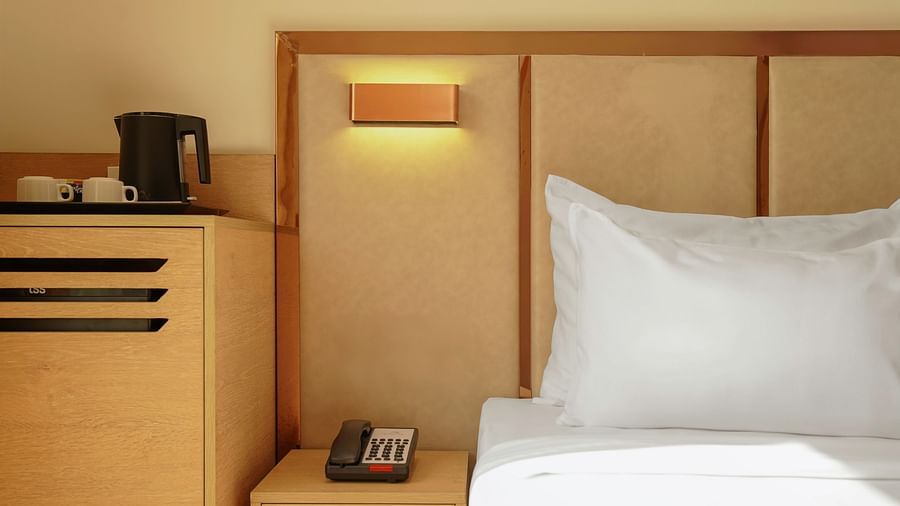 A hotel nightstand with a landline telephone, kettle, and cups. A bed with white linens is in the background - 