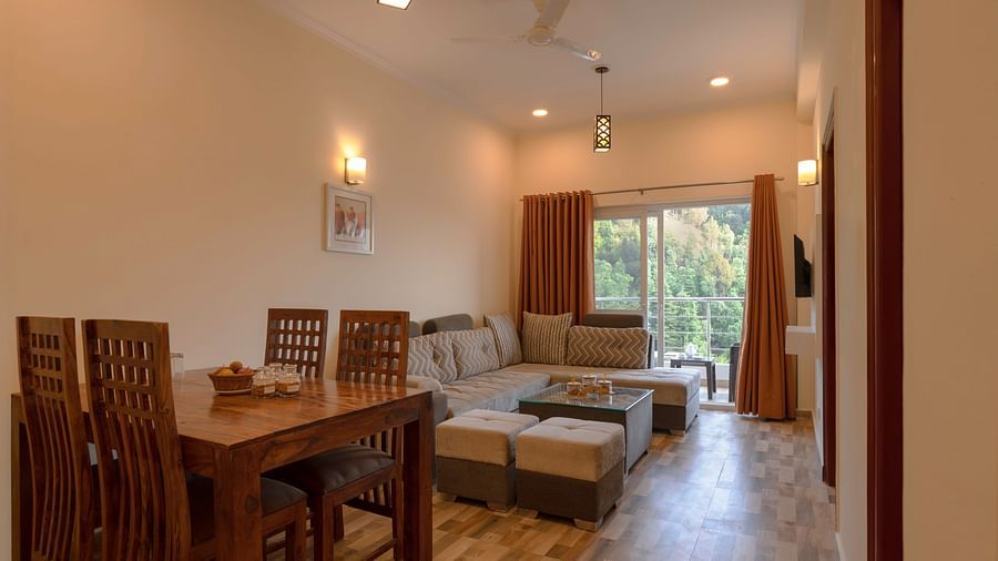 living and dining area includes a wooden table with chairs, a corner sofa, pouffes, and a window with orange curtains overlooking greenery at Clarks Inn, Near Kainchi Dham.
