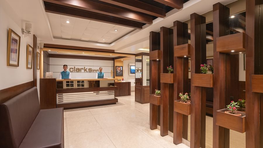 The reception area of Clarks Inn, Near Kainchi Dham shows two staff members behind a counter, with seating and decorative wooden pillars with plants.