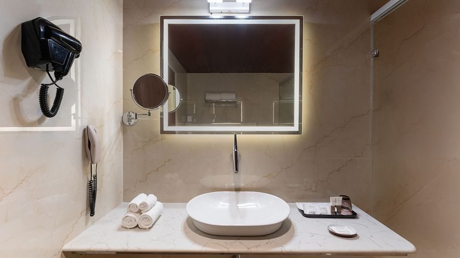 Modern hotel bathroom with an illuminated mirror, bowl-style sink, towels, mounted hairdryer, and a sleek, beige wall at Clarks Inn Suites, Hyderabad