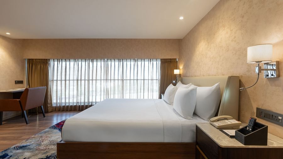 Side angle of a neatly made king-size bed with soft pillows, bedside lamps, and writing desk placed beside full-length curtains at Clarks Inn Suites, Hyderabad