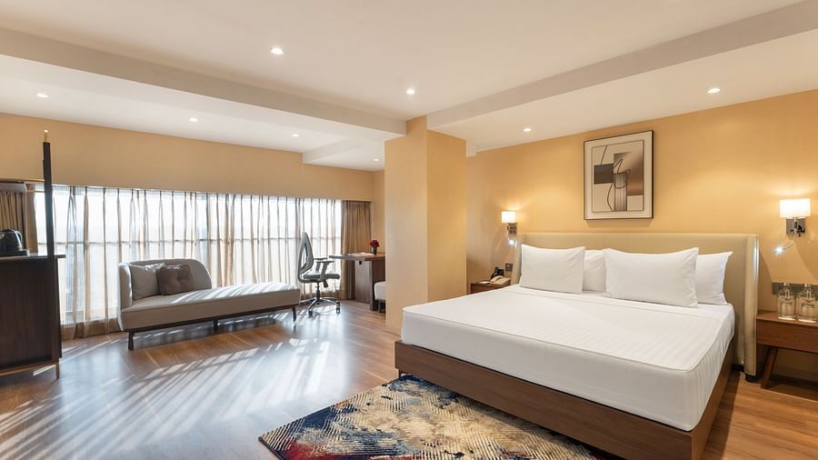 Wide-angle view of a premium suite with a cosy king bed, rug, lounge seating, and abundant natural light from large windows at Clarks Inn Suites, Hyderabad