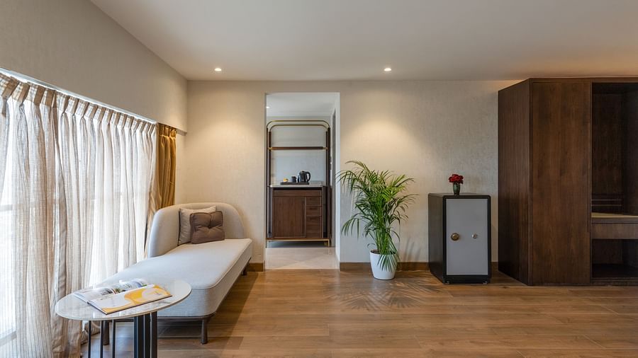Stylish lounge zone with light wood floors, potted plant, chaise, mini fridge, and view towards the bathroom area at Clarks Inn Suites, Hyderabad
