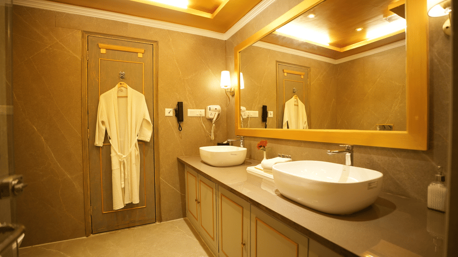 An Executive Suite bathroom at Clarks Khajuraho featuring a vanity with a sink, a large mirror, bathrobes hanging, and a wooden door entrance.