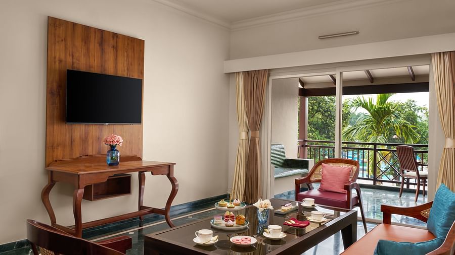 The living room in Executive Suite, featuring a comfortable seating area, a flat-screen TV, and a balcony overlooking a swimming pool and lush greenery at DGV Resort by Treat, Silvassa