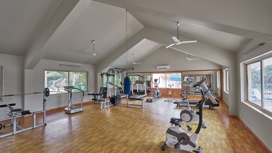 The gym at DGV Resort by Treat, Silvassa with wooden flooring, modern fitness equipment, large mirrors, ceiling fans, and windows offering natural light and garden views