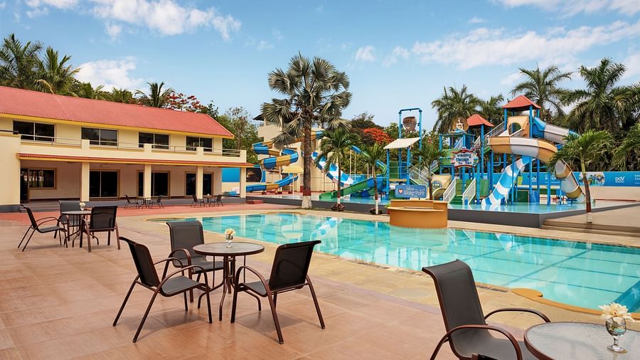 A swimming pool at DGV Resort by Treat, Silvassa with colourful slides, splash structures, a swimming pool, poolside seating, palm trees, and a building with red-tiled roof.