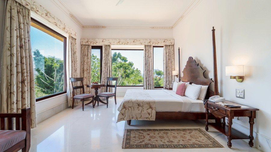 A spacious bedroom featuring a dark wood 2-poster bed, elegant drapery, and large windows with garden views - Fateh Garh, Udaipur