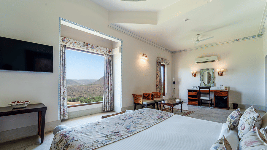 A bright guest room with a mounted television, seating area, and a view of the rugged Udaipur landscape - Fateh Garh, Udaipur