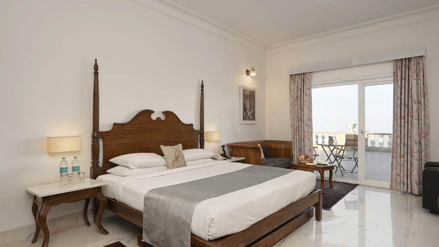 Deluxe Room with Balcony at Fateh Safari Resort, Kumbhalgarh featuring a double size bed, bedside table with water bottles, a seating area, and a balcony attached which provides an outdoor vide.