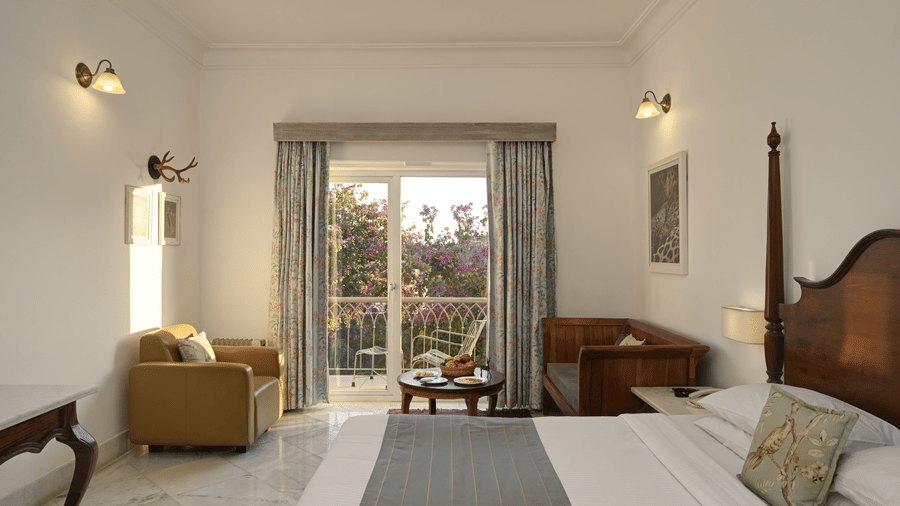 Deluxe Room with Balcony at Fateh Safari Resort, Kumbhalgarh featuring a seating area with sofa sets and centre table, a duble size bed, and a balcony attached which have curtains parted during the day.