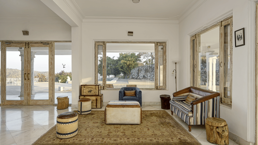 A lobby at Fateh Safari Resort, Kumbhalgarh featuring wooden sofa set, stools, and centre table and windows with outdoor views.