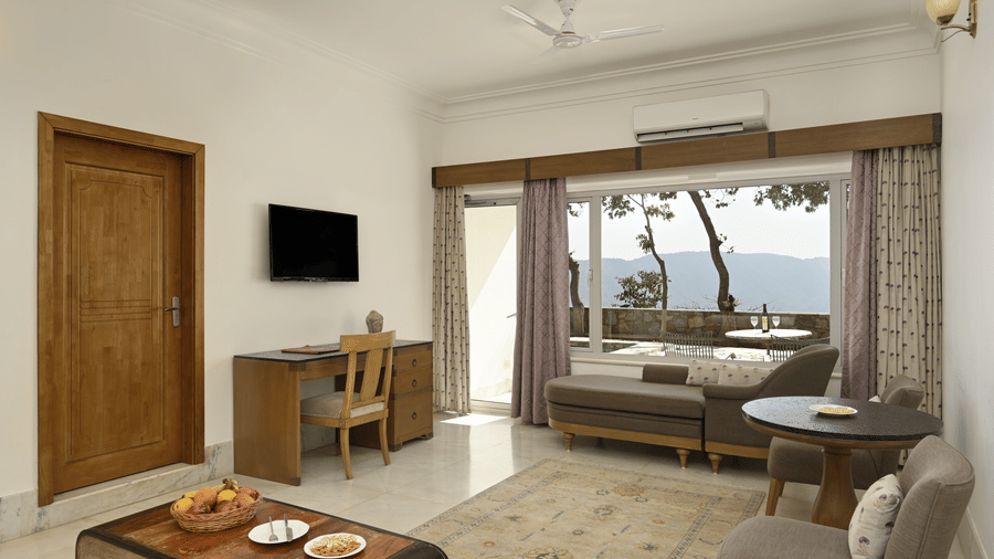 Plunge Pool Suite at Fateh Safari Suites featuring seating space with sofa set, centre tables, workstation with desk and chair, flat TV screen, and a view of the hill from the window.