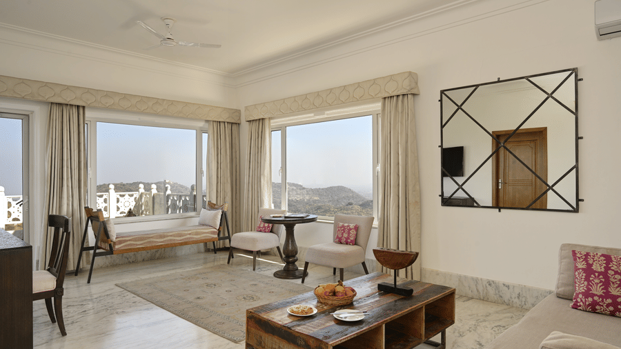 Plunge Pool Suite at Fateh Safari Suites featuring a seating area with sofa chairs, centre tables, work desk and chair, mirror on the wall, and windows which provides views of green hills.