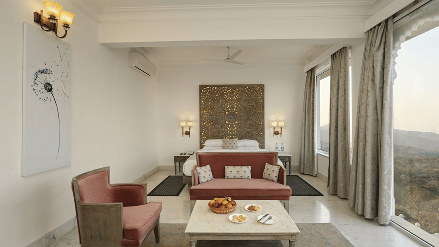 Seating area of Plunge Pool Suite in Fateh Safari Suites featuring sofa sets, a centre table with fruit basket, cookies, and other items, bedside tables on either side, and a huge window with view of greenery.