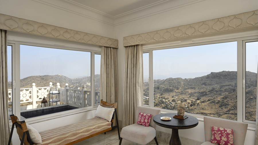 Large windows of Plunge Pool Suite in Fateh Safari Suites offering the views of green hills. Inside the room there are seating areas with sofa set and a centre table.