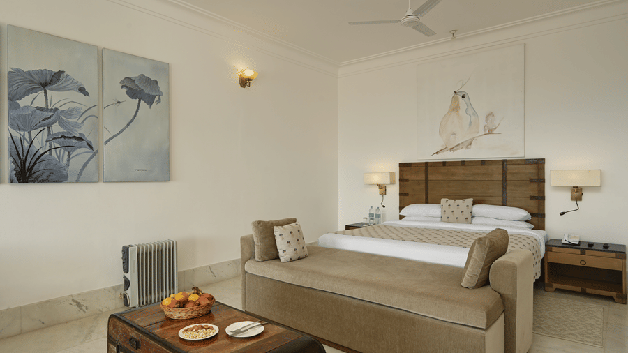 Plunge Pool Suite with Jacuzzi at Fateh Safari Suites, Kumbhalgarh featuring a bed with beside tables and lamps, a sofa at the centre of the room, a table with fruits, and a trolley bag at one corner.