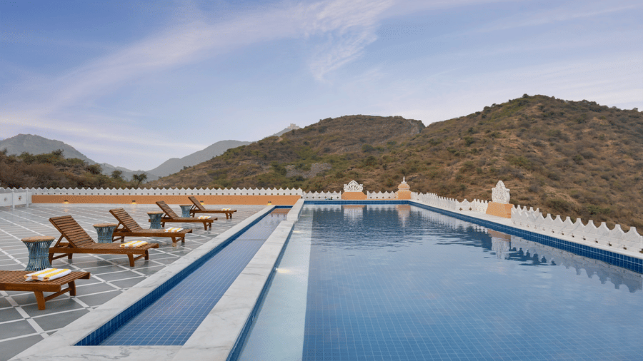 Swimming pool with loungers next to it and mountains in the background at Fateh Vilas Udaipur. Get access to the contact of Fateh Vilas by Fateh Collection for more information.