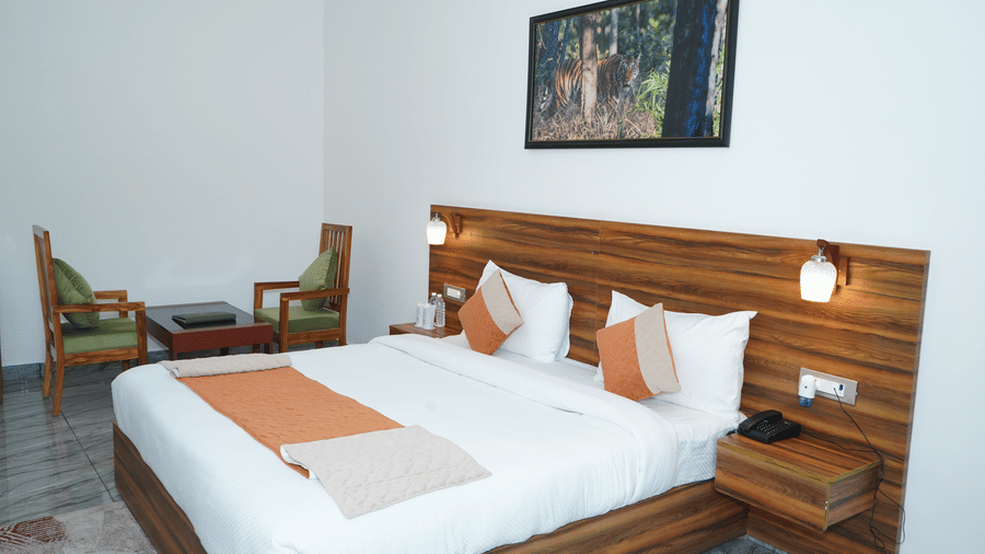 Well-lit guest bedroom featuring a king size bed with crisp white linens, a wooden panel headboard with warm reading lamps, framed wildlife wall art, and a sitting area - Five Elements Hotels, Corbett Crown Resort, Jim Corbett
