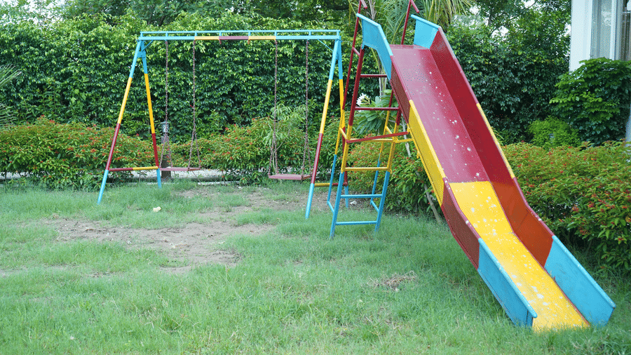 Children's outdoor playground area on a grass lawn featuring a green swing set and a red, yellow, and blue plastic slide - Five Elements Hotels, Corbett Crown Resort, Jim Corbett