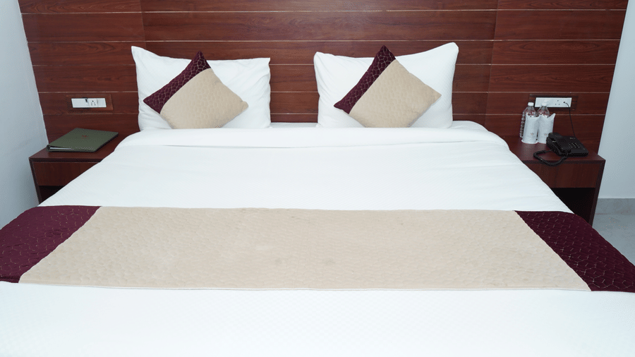 Frontal view of a king sized bed neatly made with white linens, decorative bed runner, and plush accent pillows against a dark wood headboard - Five Elements Hotels, Corbett Crown Resort, Jim Corbett