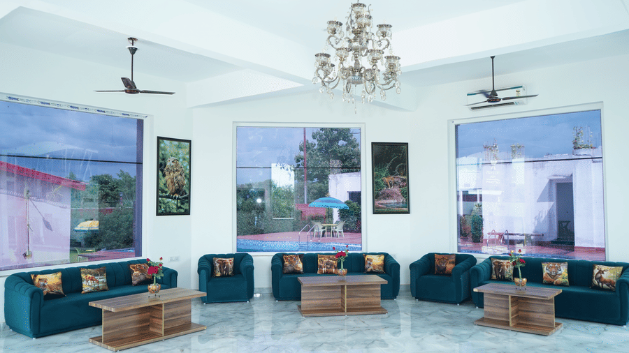 Spacious and bright resort lobby lounge area with plush green sofas, coffee tables, large glass windows, ceiling fans, and a chandelier - Five Elements Hotels, Corbett Crown Resort, Jim Corbett