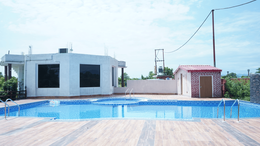 Outdoor rectangular swimming pool surrounded by a light brown paved pool deck, featuring an adjacent small structure and blue skies under clear sunny weather - Five Elements Hotels, Corbett Crown Resort, Jim Corbett