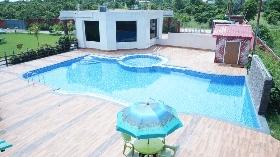 Elevated high-angle view of the outdoor swimming pool area showing the crystal blue water, paved patio area, and a blue patio table umbrella - Five Elements Hotels, Corbett Crown Resort, Jim Corbett