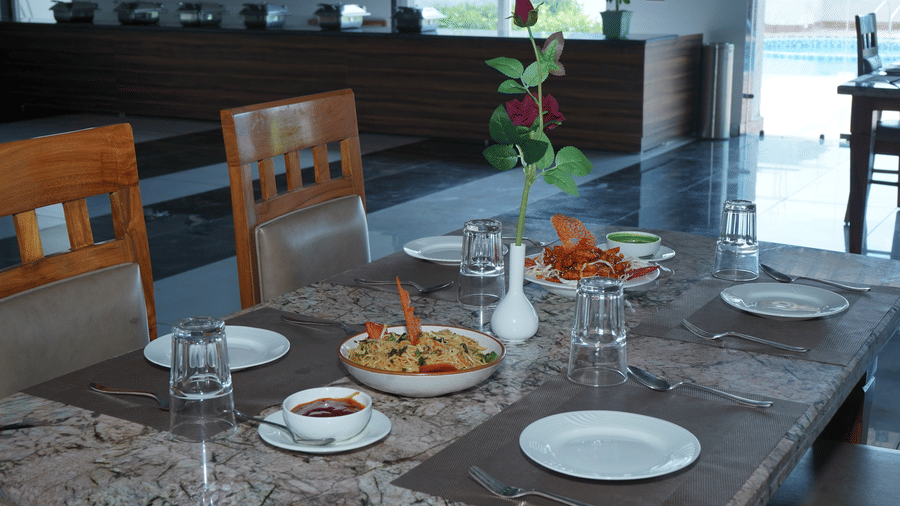 Indoor dining room setup featuring a wooden dining table arranged with plates, water glasses, serving bowls with cooked food, and a red rose centrepiece - Five Elements Hotels, Corbett Crown Resort, Jim Corbett