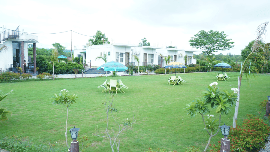 Scenic wide view of single-story resort cottages nestled behind a spacious green garden lawn adorned with small trees and outdoor table seating - Five Elements Hotels, Corbett Crown Resort, Jim Corbett