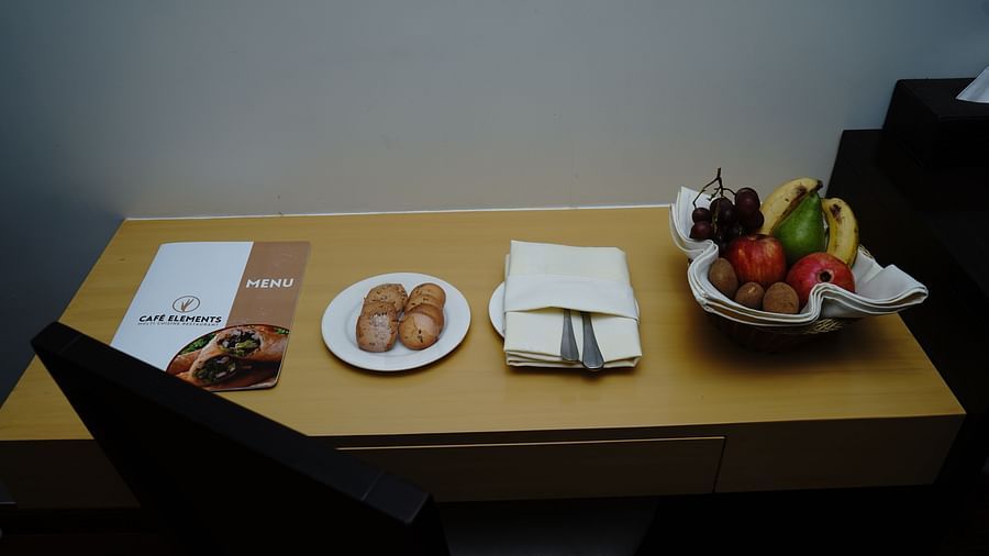 An image of a chair and desk where a menu book,  cookies, cutlery, and fruit basket is placed at Five Elements Hotels North Avenue, Delhi