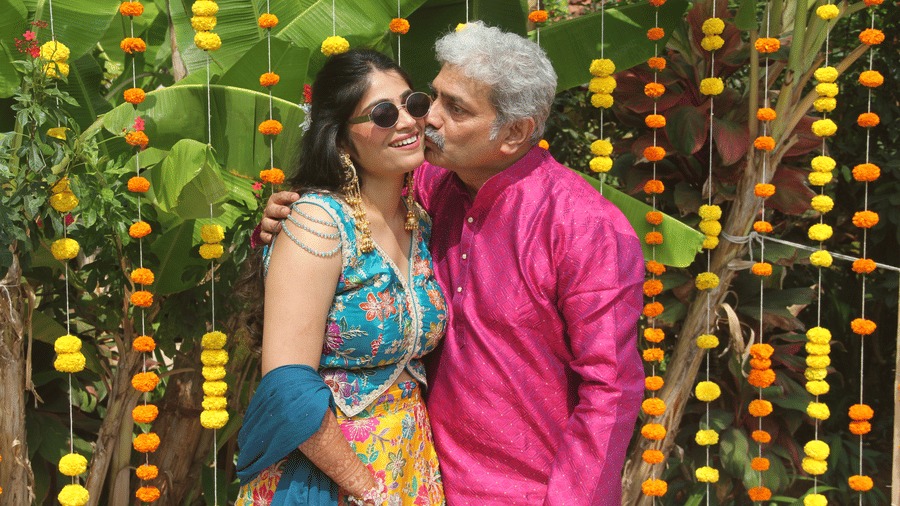 Man in a pink kurta kissing a smiling woman in a blue and yellow outfit on the cheek before a backdrop of hanging marigold strings and banana leaves.