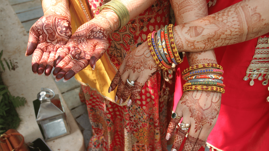 Close-up of 3 women's hands and forearms decorated with intricate henna patterns, wearing colorful bangles, pearl rings, and gemstone rings.