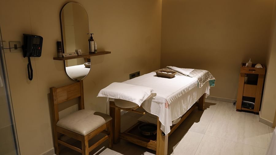 Massage room with a wooden bed covered in white linens and a folded patterned blanket, a chair beside a wall-mounted mirror and shelf with bottles, a wall telephone, and a wooden trolley in the corner under soft ambient lighting - Golden Tulip, Lucknow