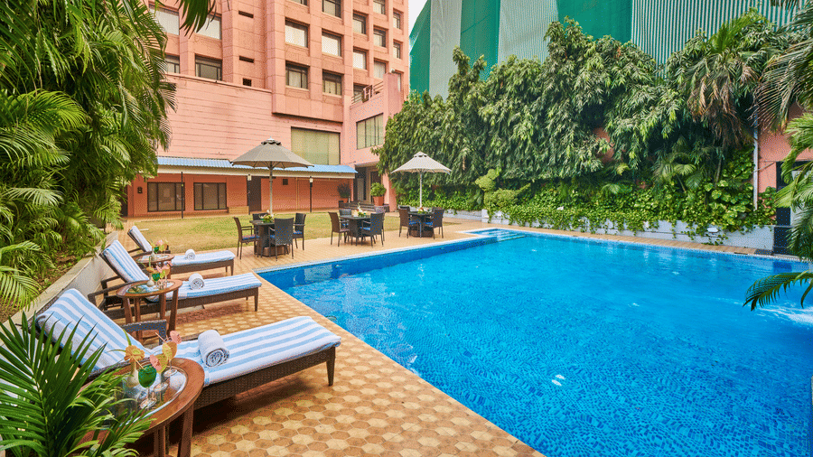 An outdoor swimming pool during the day with blue water, lounge chairs, and a multi-story building in the background | HHI Varanasi