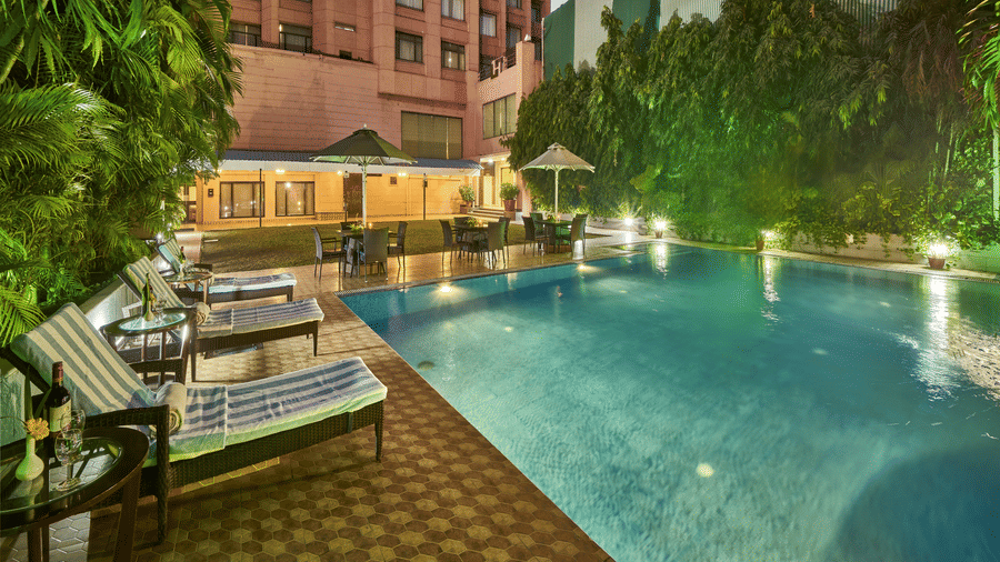 An outdoor swimming pool at night with lounge chairs on a tiled deck and trees in the background | HHI Varanasi