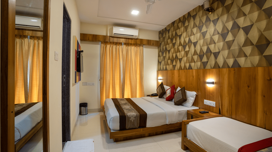 A bedroom at Home2 India with 2 beds, patterned wall panel, wooden accents, bedside lighting, a window with curtains, and a mirror near the entrance.