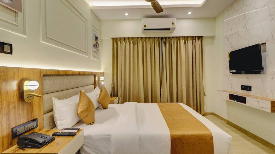 A bedroom at Home2 India with a king size bed, pillows, bedside table, wall-mounted TV, air conditioner, curtains, ceiling fan, and soft lighting.