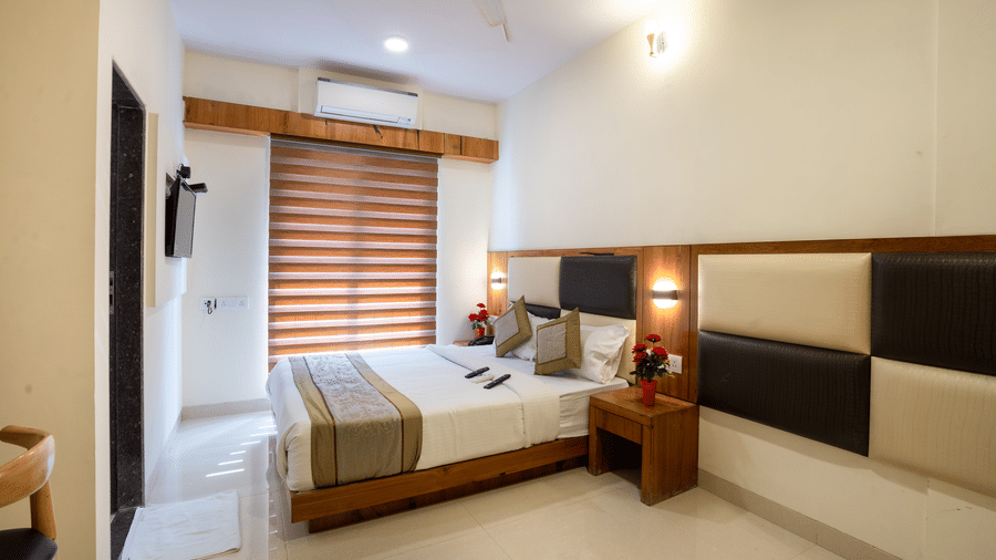 A bedroom at Home2 India with a double bed, pillows, bedside table, wall lights, window with blinds, wall paneling, and a door visible on one side