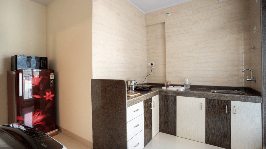 A kitchen area at Home2 India with countertop, sink, cabinets, drawers, tiled walls, a refrigerator, and part of a washing machine visible in the foreground.